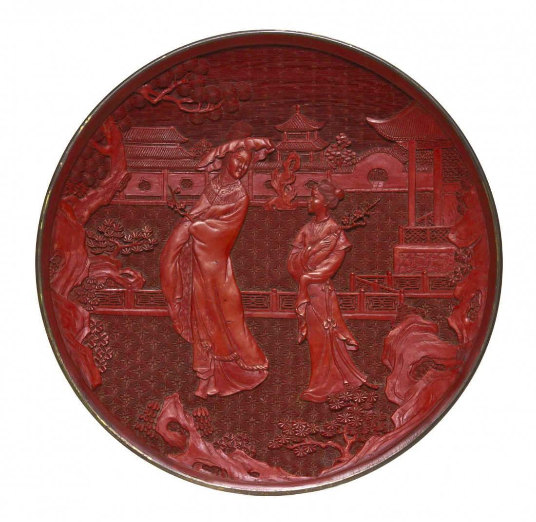 A Carved Cinnabar Lacquer Tray, Diameter 10 inches: A Carved Cinnabar Lacquer Tray, having decoration of a woman and a young girl walking in a garden holding flowering branches, having pagodas and trees in the background. Diameter 10 inches.