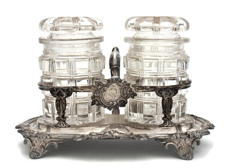 An English Silverplate and Cut Glass Canister Set,: An English Silverplate and Cut Glass Canister Set, having two cylindrical canisters within the handled silverplate base with scrolling decoration and feet. Height overall 5 1/2 inches.