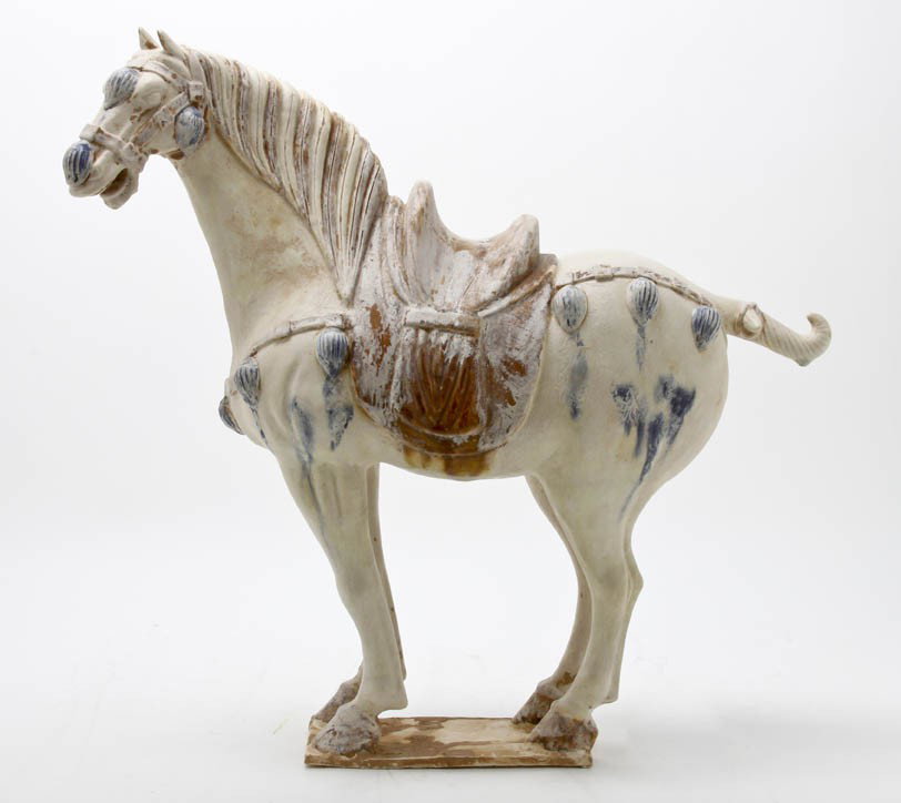 A Tang Style Horse, Height 19 inches.: A Tang Style Horse, in standing pose with lowered head, the body having a white iridescent white glaze inclusions, the brown saddle and bridal decorated with blue tassels. Height 19 inches.