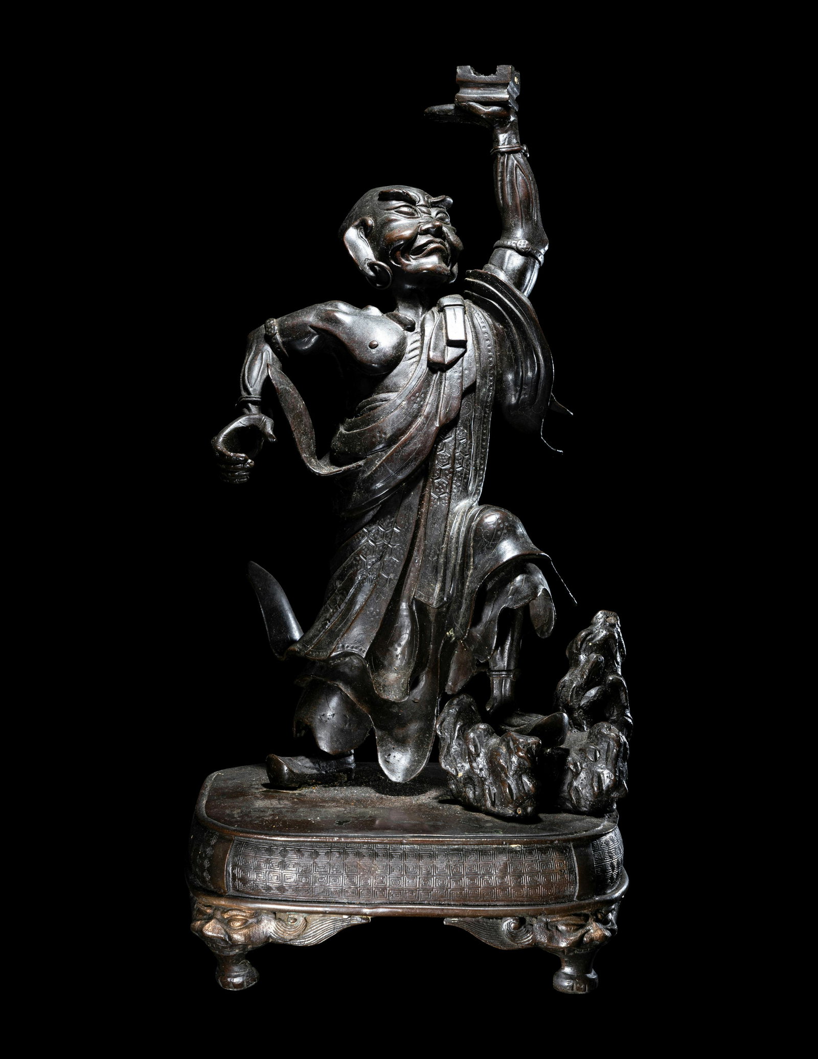 A Japanese Bronze Figure of Daruma: A Japanese Bronze Figure of Daruma Late Meiji Period the figure cast standing, facing upwards, his left hand raised above his head, holding a stand, the right arm stretched out, the left leg resting o