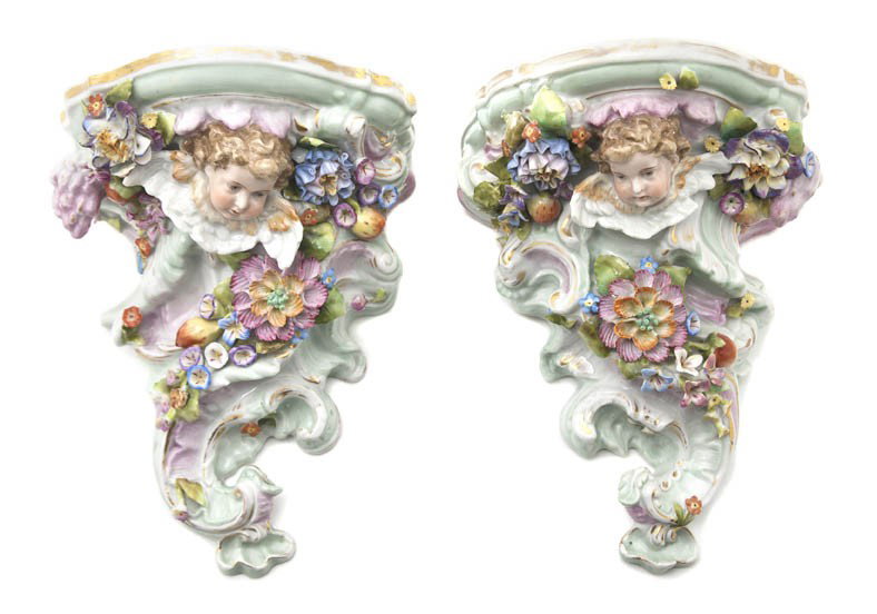 A Pair of Continental Porcelain Wall Brackets, Hei: A Pair of Continental Porcelain Wall Brackets, each having a winged putto mask and applied flowers upon scrolling supports, with gilt highlights throughout. Height 7 7/8 inches.