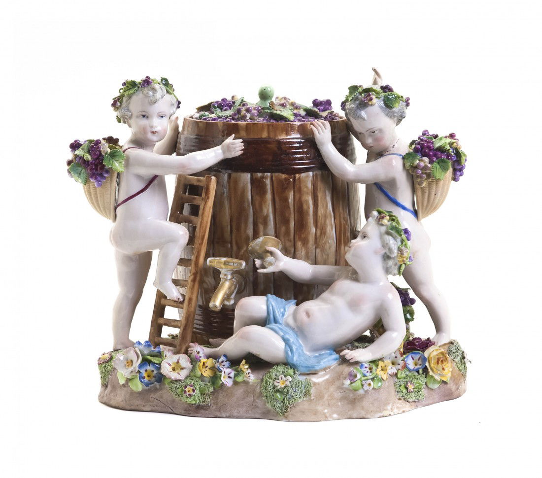 A Meissen Porcelain Biscuit Jar, Width 10 5/8 inch: A Meissen Porcelain Biscuit Jar, depicting three putti before a wine barrel with baskets of grapes, set on a naturalistic base with applied flowers and foliage, having dotted and crossed swords mark i