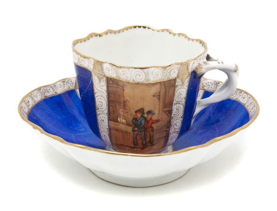 A Meissen Porcelain Cup and Saucer, Width of wide: A Meissen Porcelain Cup and Saucer, each of oval form, decorated with alternating panels depicting figures in an interior over cobalt blue with faux damage, having crossed swords mark in underglaze bl