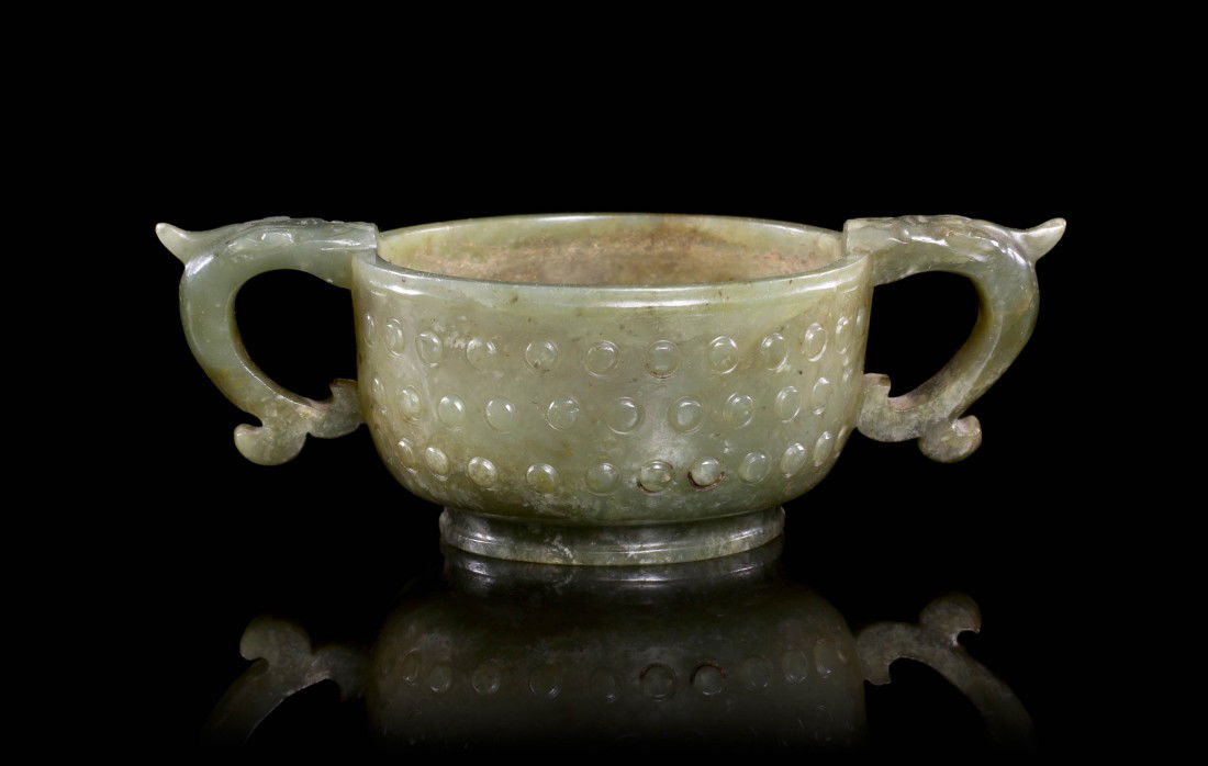 A Spinach Jade Libation Cup, Width over handles 4: A Spinach Jade Libation Cup, of circular form having raised circular bosses throughout the exterior of the bowl, with beast headed c-form handles, raised on a circular foot ring. Width over handles 4