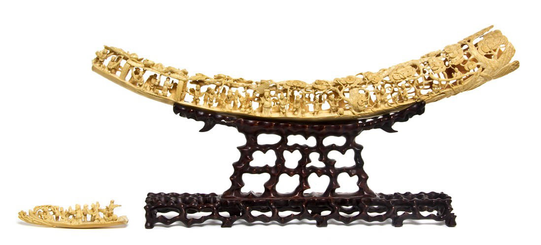 A Chinese Carved Ivory Tusk, Approximate length of: A Chinese Carved Ivory Tusk, intricately carved with figures at various activities, with craggy pine trees and massive lotus plants carved throughout, raised upon a pierce carved wood base. Approximat