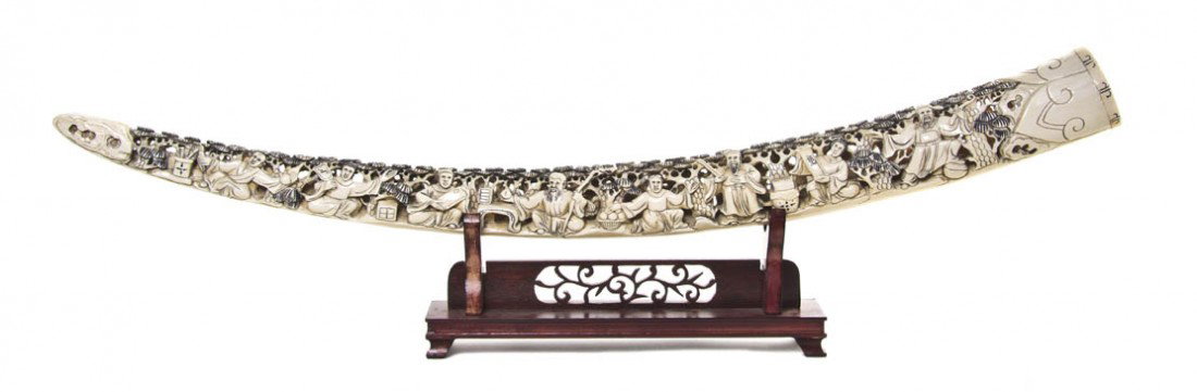 A Chinese Pierce Carved Elephant Tusk, Length of t: A Chinese Pierce Carved Elephant Tusk, having pierce carved decoration throughout, each side depicting eight immortals below pine trees, raised atop a wood stand. Length of tusk 32 inches. Property fr