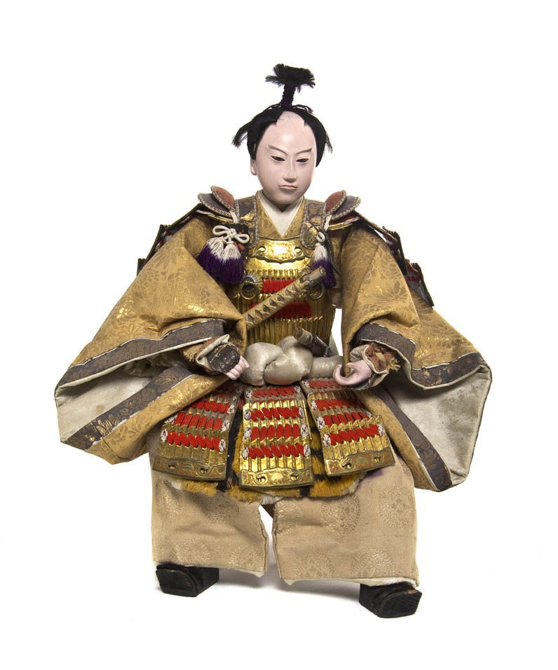 A Japanese Model of a Samurai, Height 16 1/4 inche: A Japanese Model of a Samurai, the figure of papier-mache construction with painted features, having embroidered silk clothing and shown with a sword at his side, seated on a wooden folding chair. Hei