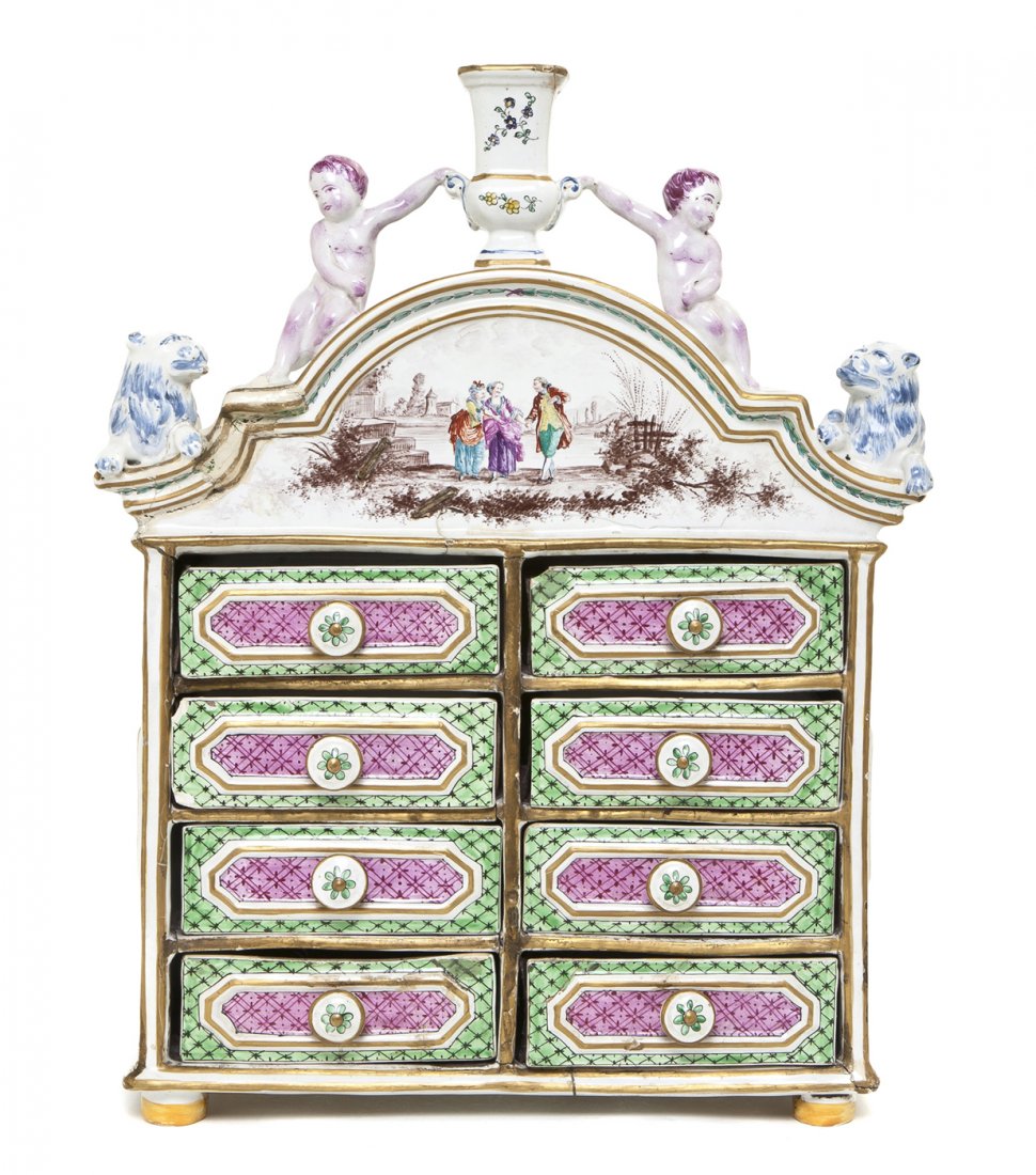 A French Faience Diminutive Chest of Drawers, Heig: A French Faience Diminutive Chest of Drawers, having an urn finial flanked by putti over the domed crest decorated with a courting scene flanked by lions, fitted with two columns of four drawers, rais