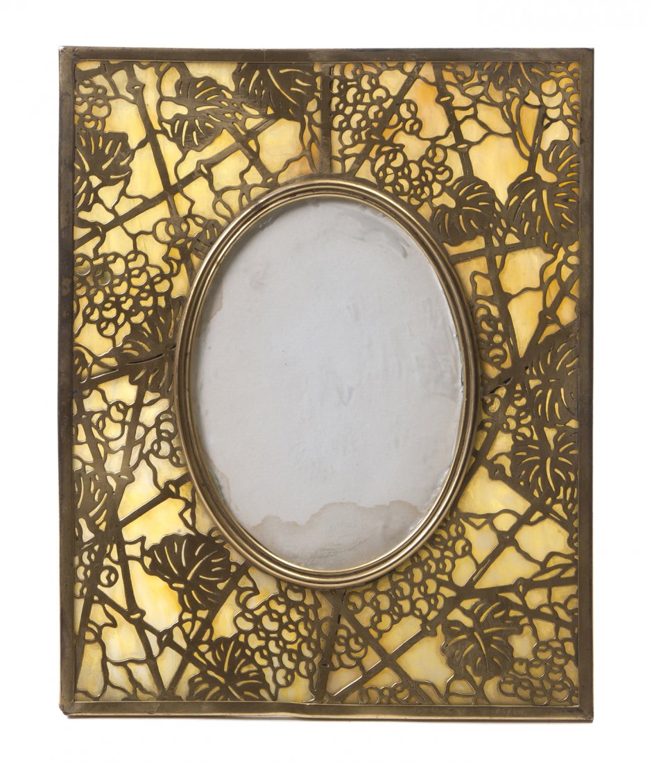 A Tiffany Studios Favrile Glass and Gilt Bronze P: A Tiffany Studios Favrile Glass and Gilt Bronze Picture Frame, in the Grapevine pattern, of vertical rectangular form with an oval opening surrounded by marbled glass, having an easel support, stamped