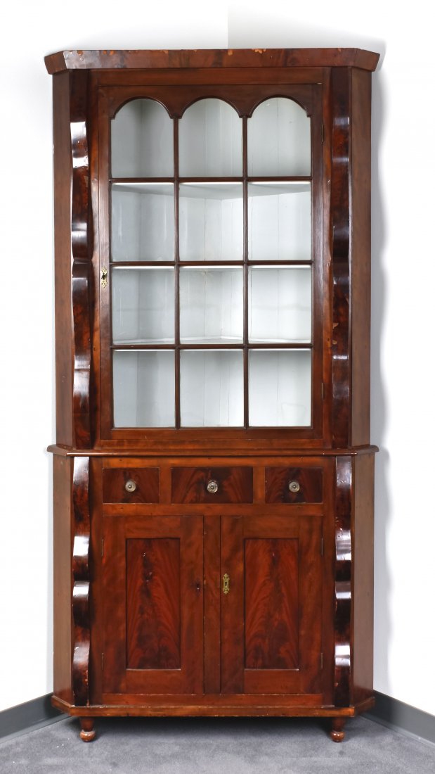 An American Empire Mahogany Veneered Corner Cabine: An American Empire Mahogany Veneered Corner Cabinet, having a flared cornice above a single glazed door flanked by molded pilasters, the base set with three short drawers above two panel doors. Height