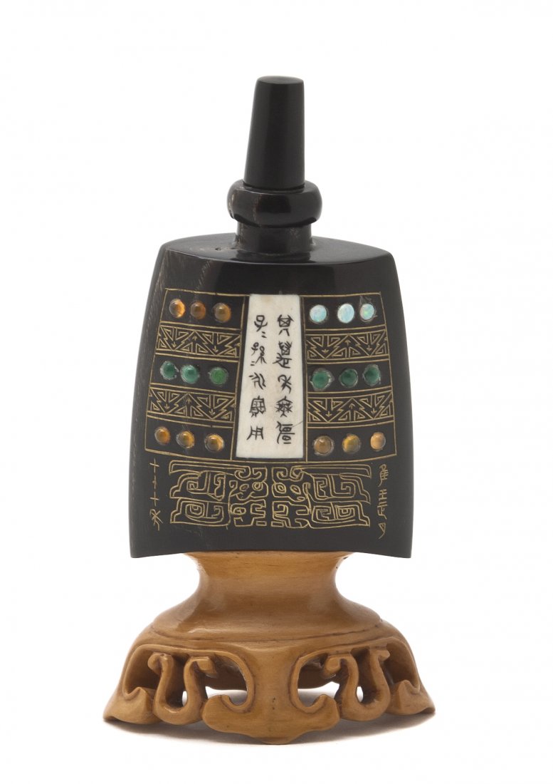A Buffalo Horn Snuff Bottle, Height overall 3 5/8: A Buffalo Horn Snuff Bottle, in the form of a Zhou dynasty bell, having inset stones, gilt inset taotie decoration and archaistic script inscriptions, mounted to a carved wood stand. Height overall 3 