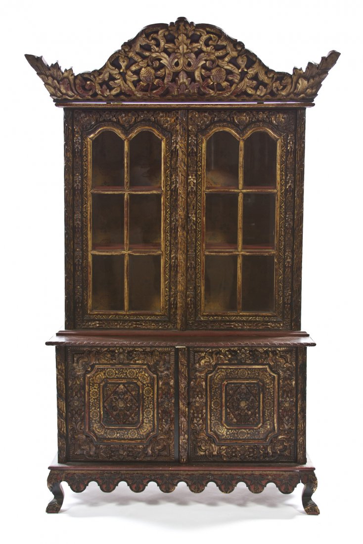 A Southeast Asian Lacquered and Parcel Gilt Cabinet: A Southeast Asian Lacquered and Parcel Gilt Cabinet, the domed projecting crest carved with leaves and scrolls over a pair of glazed doors opening to shelves, the base set with two carved panel doors 
