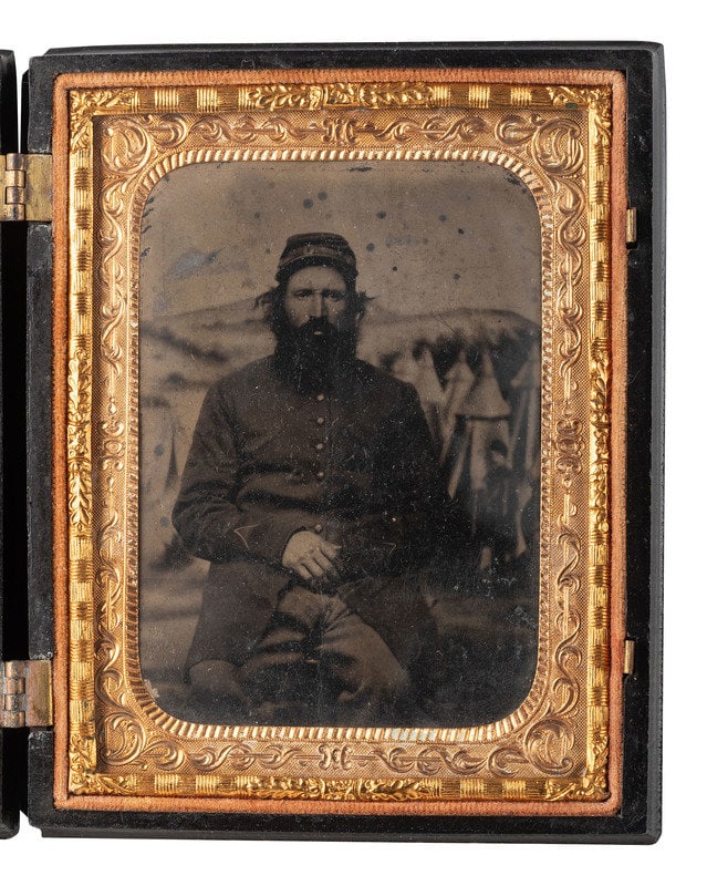 [CIVIL WAR]. Quarter plate tintype of a grizzled soldier posed before painted camp backdrop.: [CIVIL WAR]. Quarter plate tintype of a grizzled soldier posed before painted camp backdrop. Quarter plate seated portrait of a worn soldier with long sideburns, mustache, and beard, posed before a pa