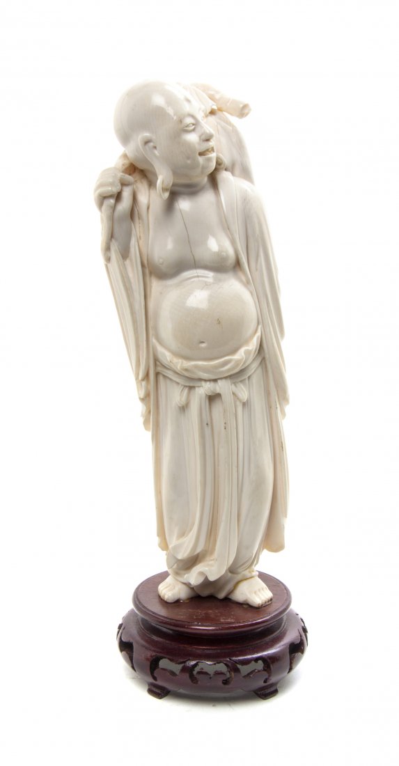A Chinese Carved Ivory Figure of a Luohan, Height overa: A Chinese Carved Ivory Figure of a Luohan, depicted standing in draped robes, his smiling face turned to the side, raised on a pierced wood base. Height overall 10 1/4 inches. Property from the Collec
