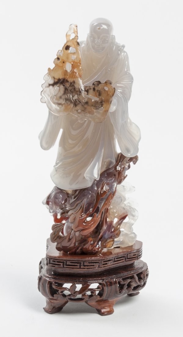 A Chinese Shadow Agate Carved Figure, Height 5 7/8 inch: A Chinese Shadow Agate Carved Figure, depicting a robed monk, standing upon a naturalistic lingzhi fungus encrusted base, further raised upon a pierce carved wood stand. Height 5 7/8 inches. Property 