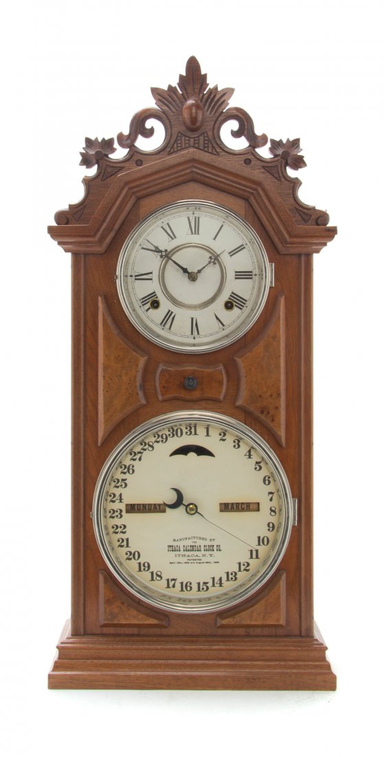 An American Double Dial Walnut Clock, Ithaca Clock Co.,: An American Double Dial Walnut Clock, Ithaca Clock Co., having a carved peak crest over the circular time dial with Roman hours and the date dial with Arabic numerals, raised on a stepped plinth base,