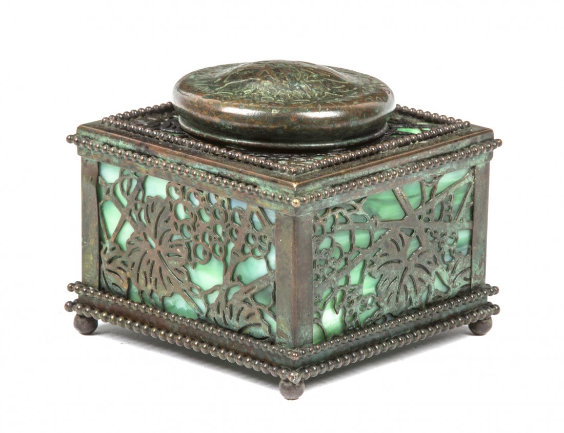 A Tiffany Studios Patinated Bronze and Favrile Glass In: A Tiffany Studios Patinated Bronze and Favrile Glass Inkwell, in the Grapevine pattern, of square form with a hinged circular lid, having a glass inkwell insert marked The Davis, with green glass inse