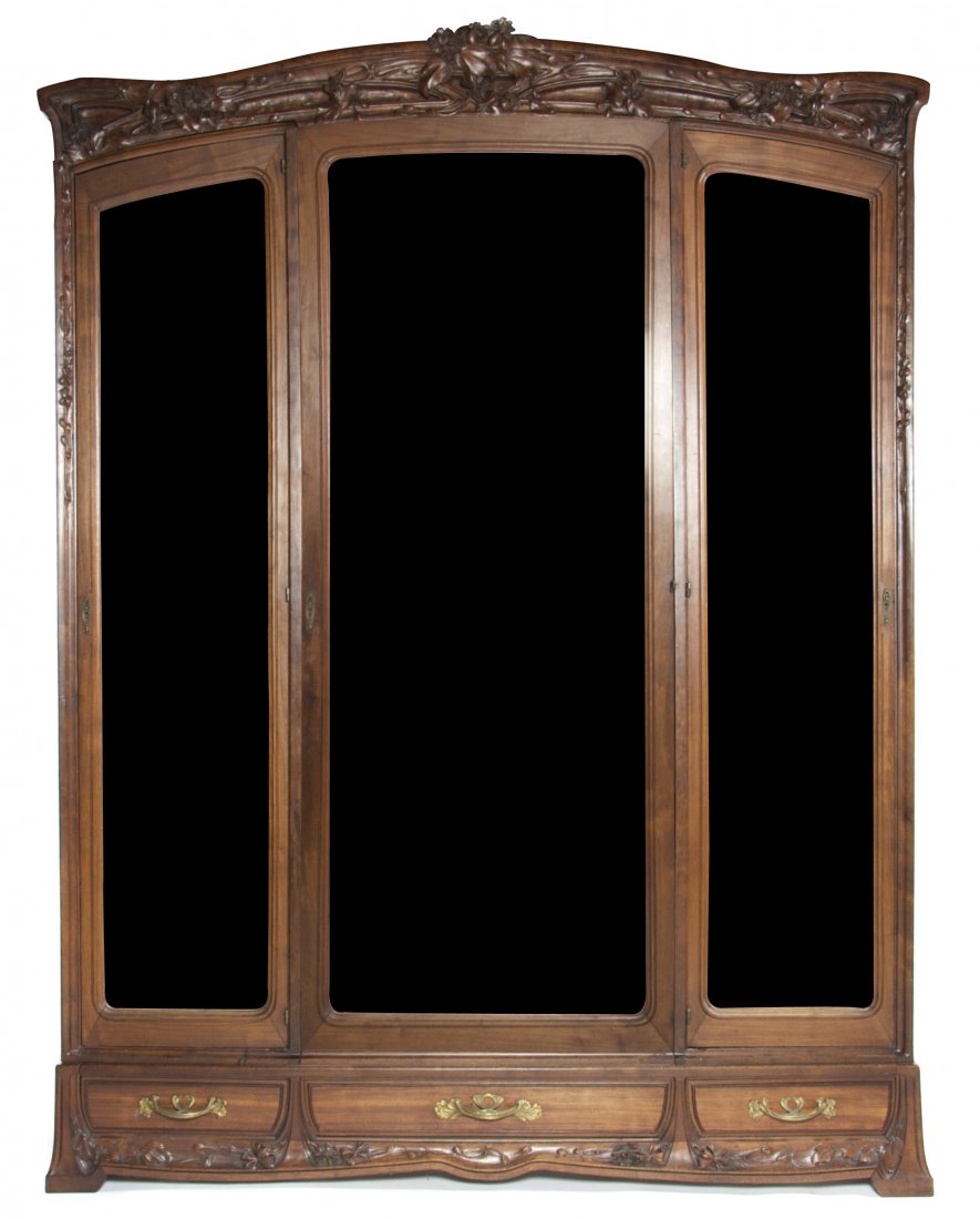 A Louis Majorelle Art Nouveau Mahogany Armoire, Height : A Louis Majorelle Art Nouveau Mahogany Armoire, having a carved arched pediment with floral decoration above a central mirrored cabinet flanked by mirrored cabinets, over three drawers, having foliate