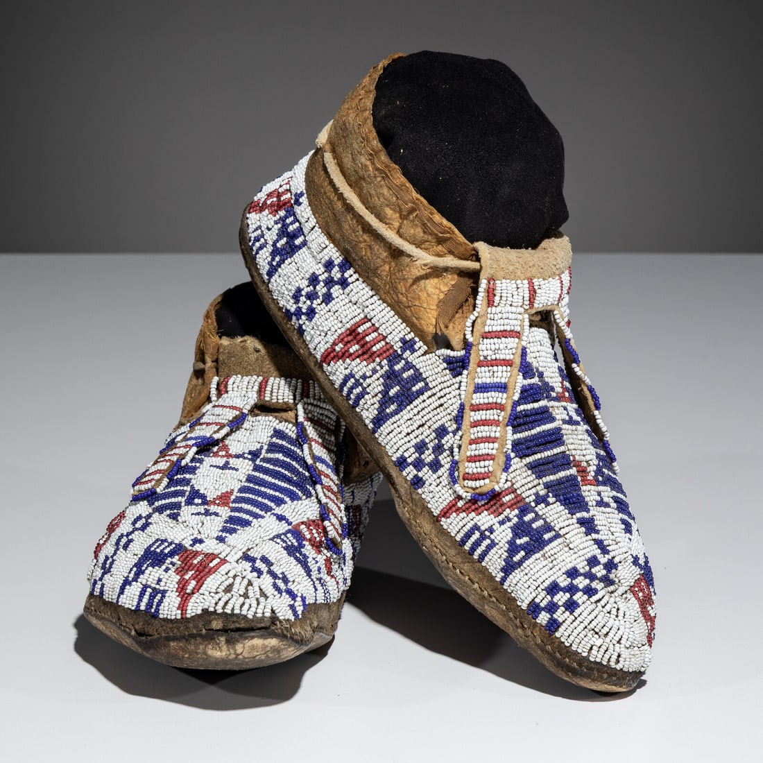 Sioux Beaded Hide Moccasins: Sioux Beaded Hide Moccasins fourth quarter 19th century sinew-sewn; with forked tongues and a beaded geometric design in colors of navy blue and red white-heart against a white ground length 10-3/4 in