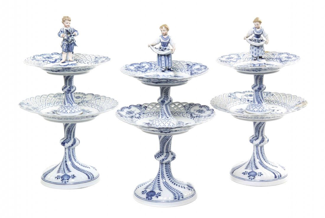 Three Meissen Porcelain Figural Pastry Stands, Height: Three Meissen Porcelain Figural Pastry Stands, each surmounted with a figure, two having a girl gathering flowers in her skirt, the other a boy with a floral wreath, each over the two graduated reticu