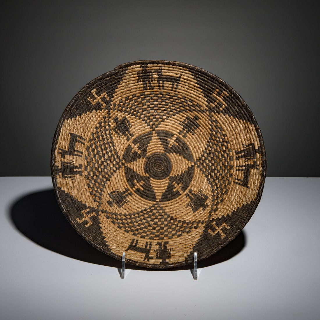 Apache Figural Basket: Apache Figural Basket early 20th century featuring eight human figures, four dogs, four whirling logs, and four crosses height 2-3/4 inches x diameter 14-1/4 inches This lot is located in Chicago.
