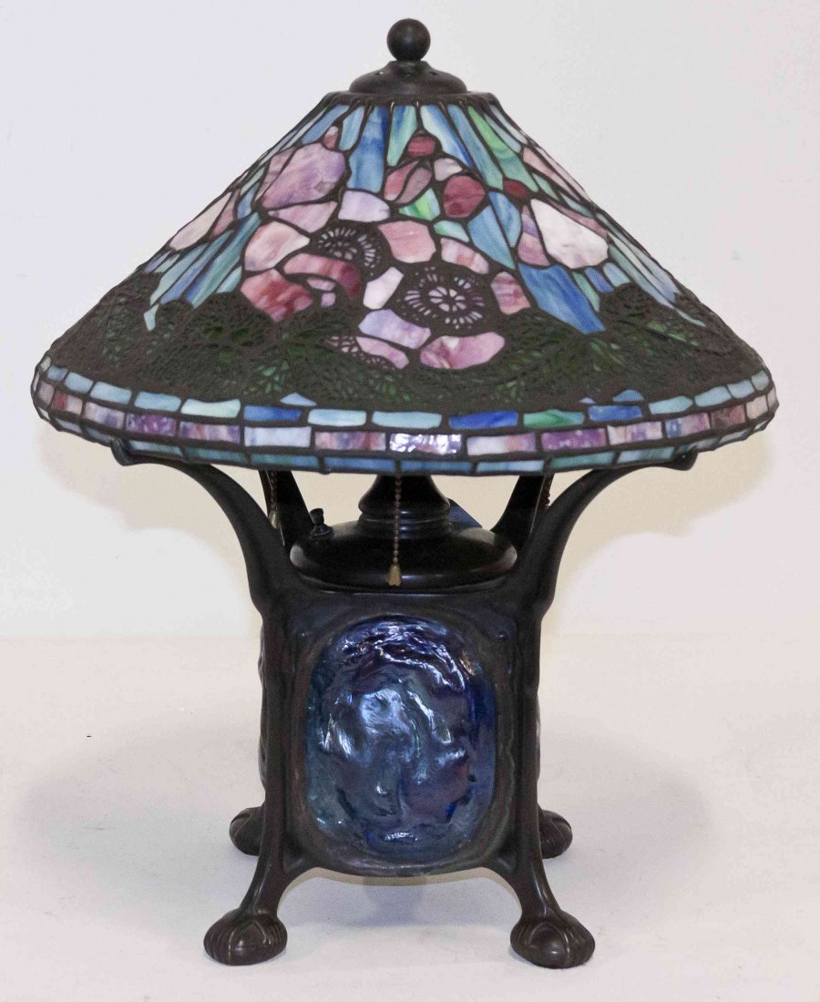 An American Leaded Glass and Bronze Table Lamp, in the: An American Leaded Glass and Bronze Table Lamp, in the manner of Tiffany Studios, the conical shade decorated with flowers above a continuous band of leaves, set on an illuminated base inset with four