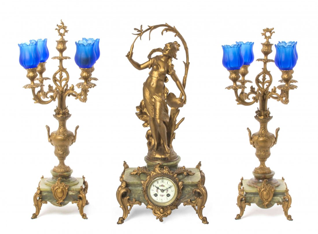 A Late Victorian Gilt Metal and Onyx Clock Garniture,: A Late Victorian Gilt Metal and Onyx Clock Garniture, after Auguste Moreau, having a circular floral painted dial with Arabic hours, within an onyx case surmounted by a gilt metal figure with an inscr