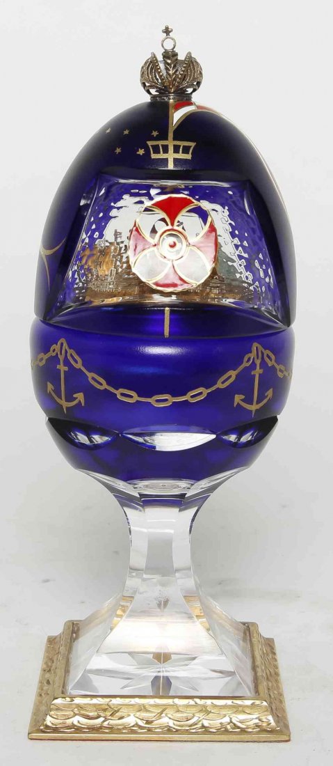 A Faberge Glass Egg, Height 8 1/4 inches.: A Faberge Glass Egg, having a crown form finial over the blue glass body opening to reveal a silver plaque with two ships, set on a faceted standard over the square foot, etched Faberge to base. Heigh