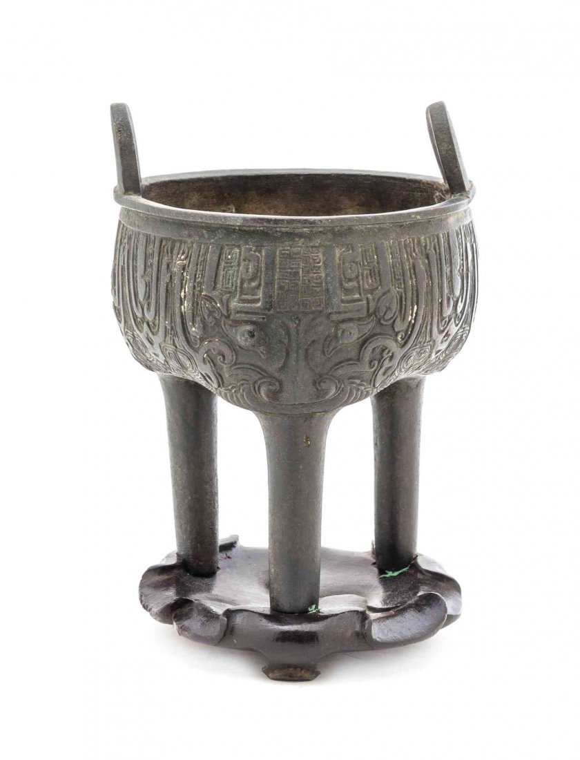 A Chinese Bronze Tripod Censer, Height 6 1/4 inches.: A Chinese Bronze Tripod Censer, having two upright loop flange handles with archaistic taotie decoration to the body, raised on a floriform hardwood stand. Height 6 1/4 inches.