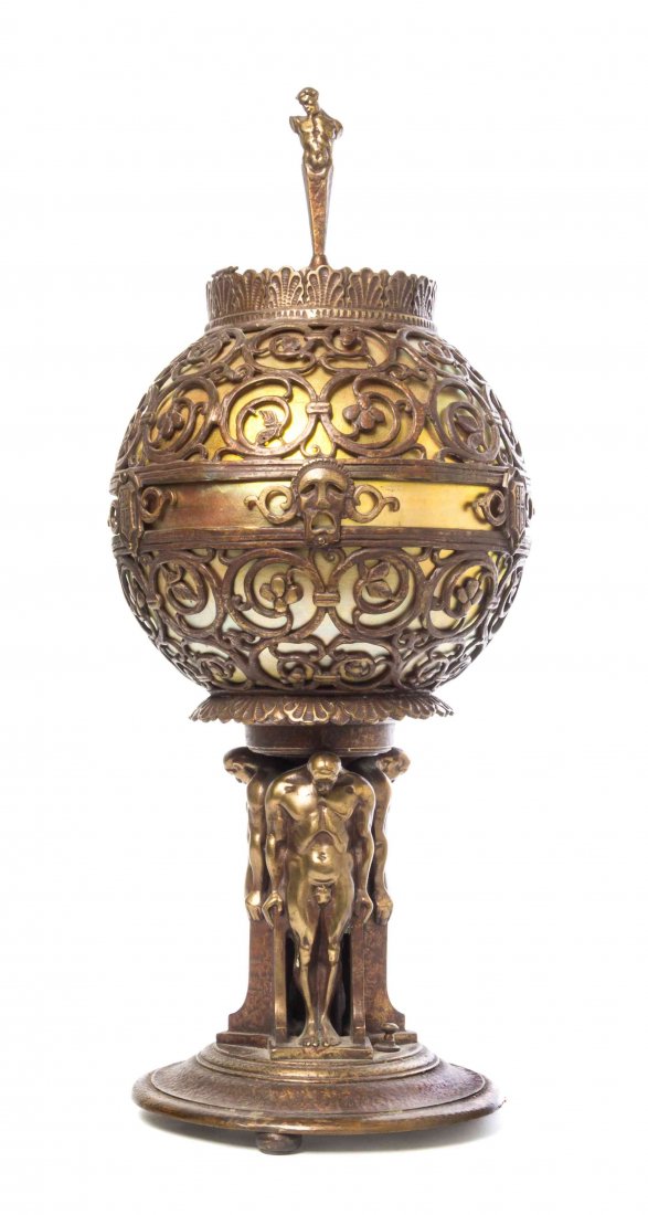 An Oscar Bach Brass Table Lamp, German/ American: An Oscar Bach Brass Table Lamp, German/ American (1884-1957), the Steuben gold Aurene glass shade of spherical form with pierced scrolling overlay, surmounted with a figural finial, the standard depic