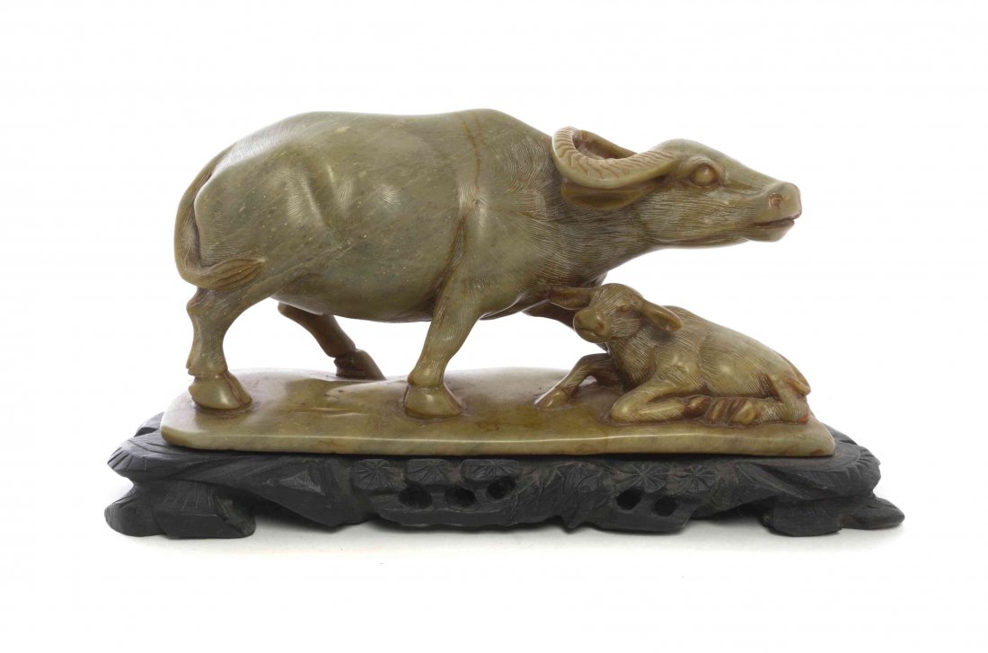 A Chinese Carved Steatite Model of a Water Buffalo,: A Chinese Carved Steatite Model of a Water Buffalo, depicting a standing water buffalo and a recumbent calf, raised on an ebonized carved wood stand depicting pine. Width 8 inches. Condition: In overa