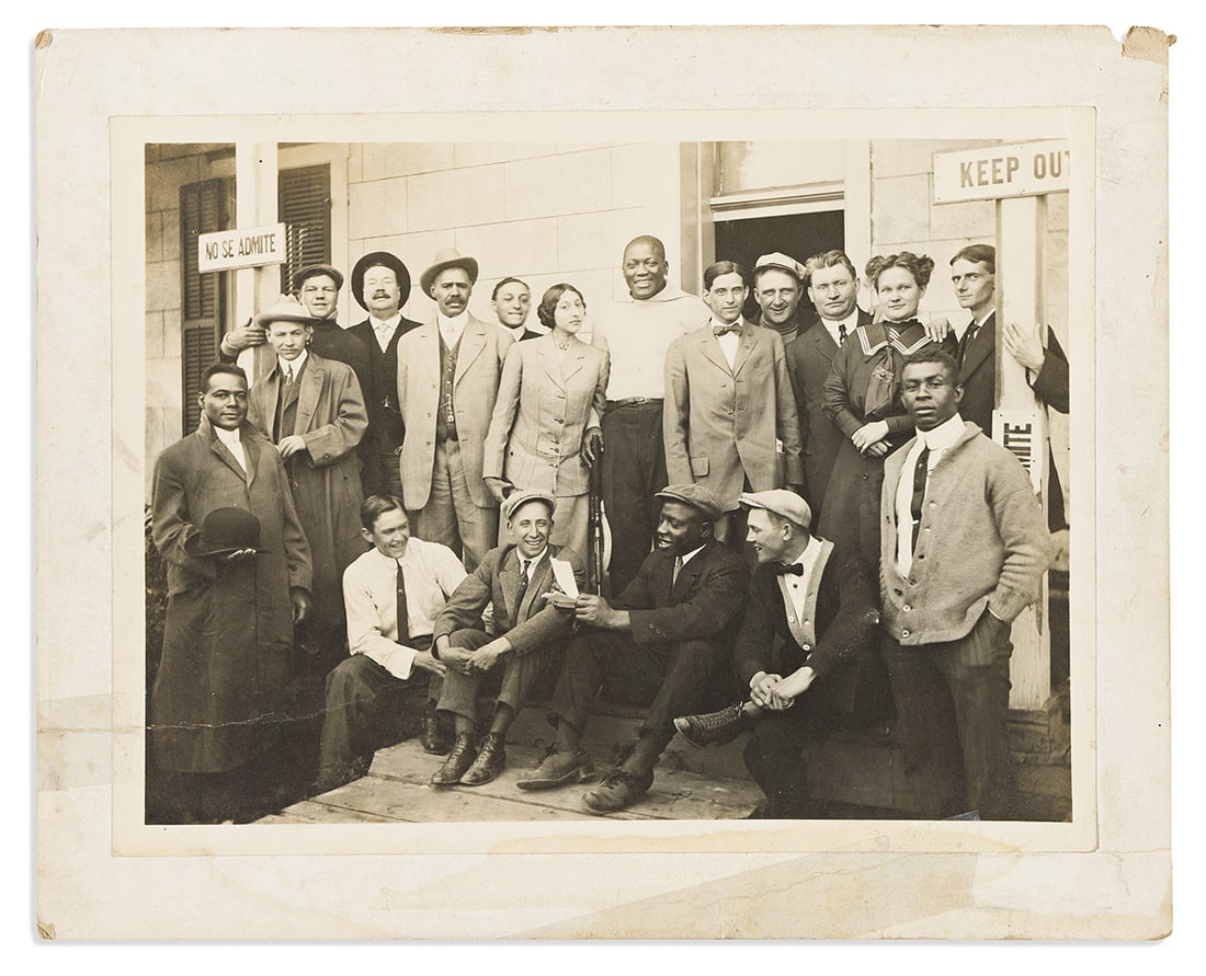 (SPORTS--BOXING.) Photograph of rifle-toting champion Jack Johnson with his entourage.: (SPORTS--BOXING.) Photograph of rifle-toting champion Jack Johnson with his entourage. Photograph, 6½ x 8½ inches, on original plain mount; two 4-inch repaired tears, modern ink caption on v