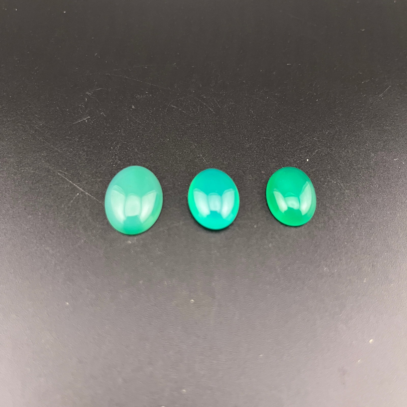 GROUP OF THREE GREEN ONYX CABOCHONS GEMSTONES: 19th Century; Long: 0.7 inch; Width: 0.5 inch; High: 0.2 inch?Long: 0.6 inch; Width: 0.5 inch; High: 0.2 inch?Long: 0.6 inch; Width: 0.45 inch; High: 0.2 inch?; Wooden base for picture and display onl