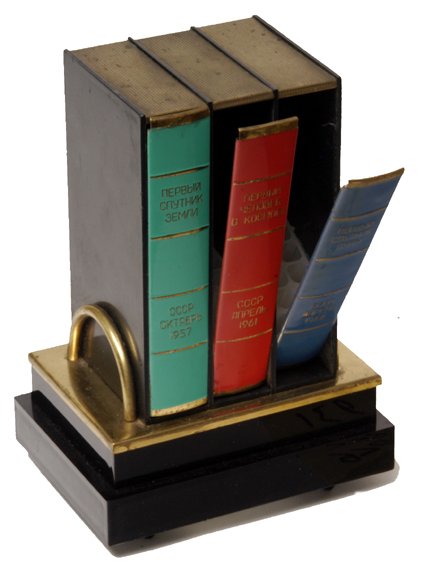 Russian Space "Cigarette / Music" Box: (4.5x3x6.5") A striking space themed Soviet cigarette music box. There are three gilt-edged "books" on a shelf. These include "First Sputnik of Earth. The USSR. October 1957" (green spine), "First Man