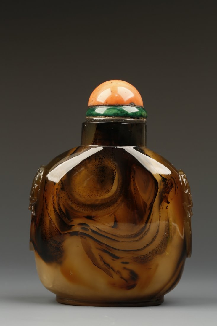 A LARGE CHALCEDONY SNUFF BOTTLE of rounded-rectangular: A LARGE CHALCEDONY SNUFF BOTTLE of rounded-rectangular form with mask-carved shoulders, the brown tones forming a swirling pattern on one side, 1800-1850, metal and coral stopper, 3.5" high Provenance