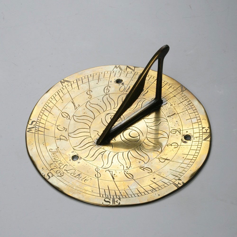 A BRASS SUN DIAL, the circular plate engraved with a ce: A BRASS SUN DIAL, the circular plate engraved with a central panel with smiling face and engraved "Tempus Labile, 1709", with pierced gnomon, 8" wide