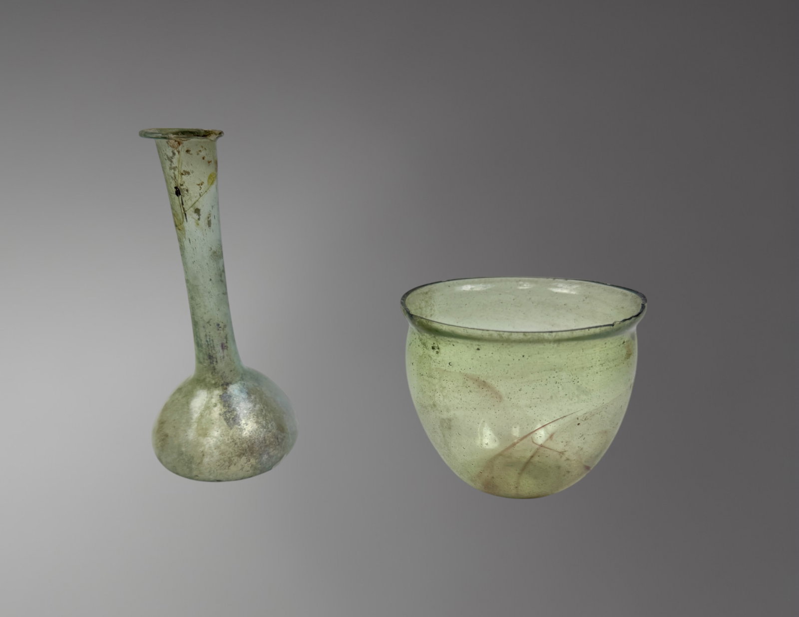 Two Roman Glass Vessels – 1st Century AD: A well-matched pair of Roman glass vessels, dating to the 1st century AD, free-blown in translucent pale blue-green glass. Both vessels feature rounded bodies with long narrow necks and slightly flare
