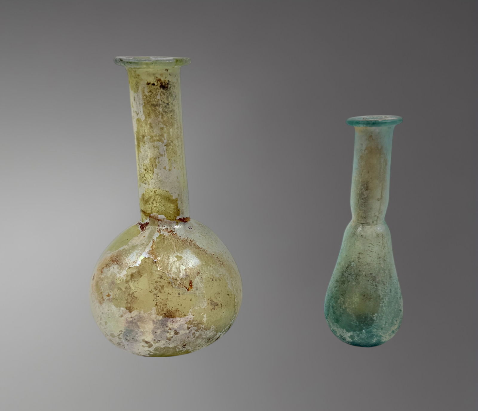 Pair of Roman Glass Vessels – 1st Century AD: A graceful pair of Roman glass vessels, dating to the 1st century AD, each free-blown in translucent pale green glass. The vessels exhibit classic forms used for storing precious liquids such as perfu