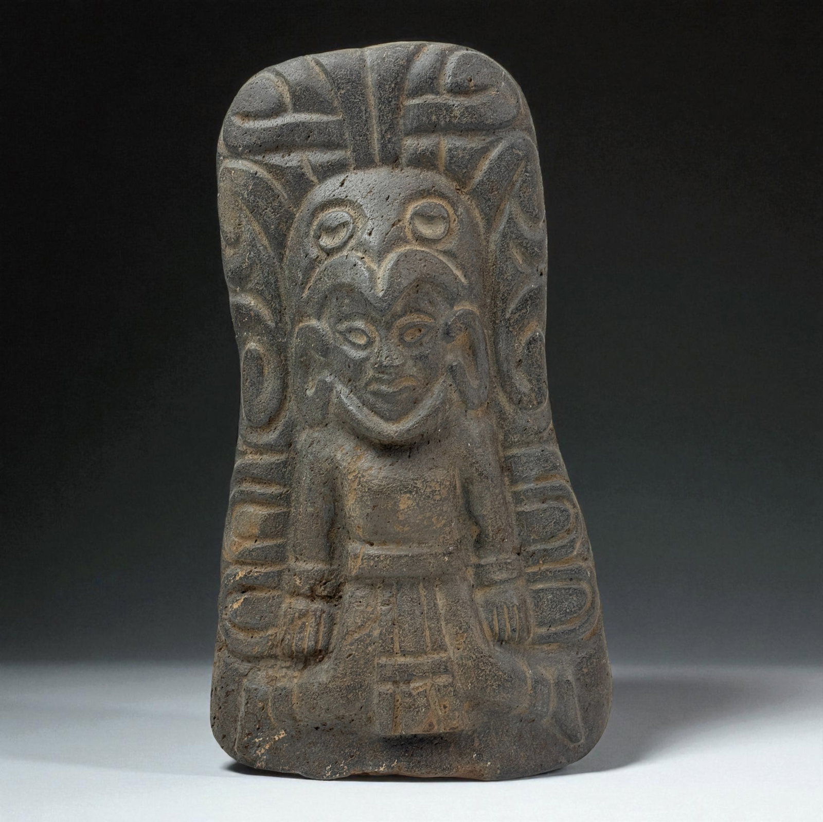 Pre-Columbian Style Basalt Deity Stela: Carved basalt stela depicting a central deity figure with elaborate headdress and geometric body detailing. Provenance: Beverly Hills Barakat Gallery Collection.13 1/2" x 7 1/2"