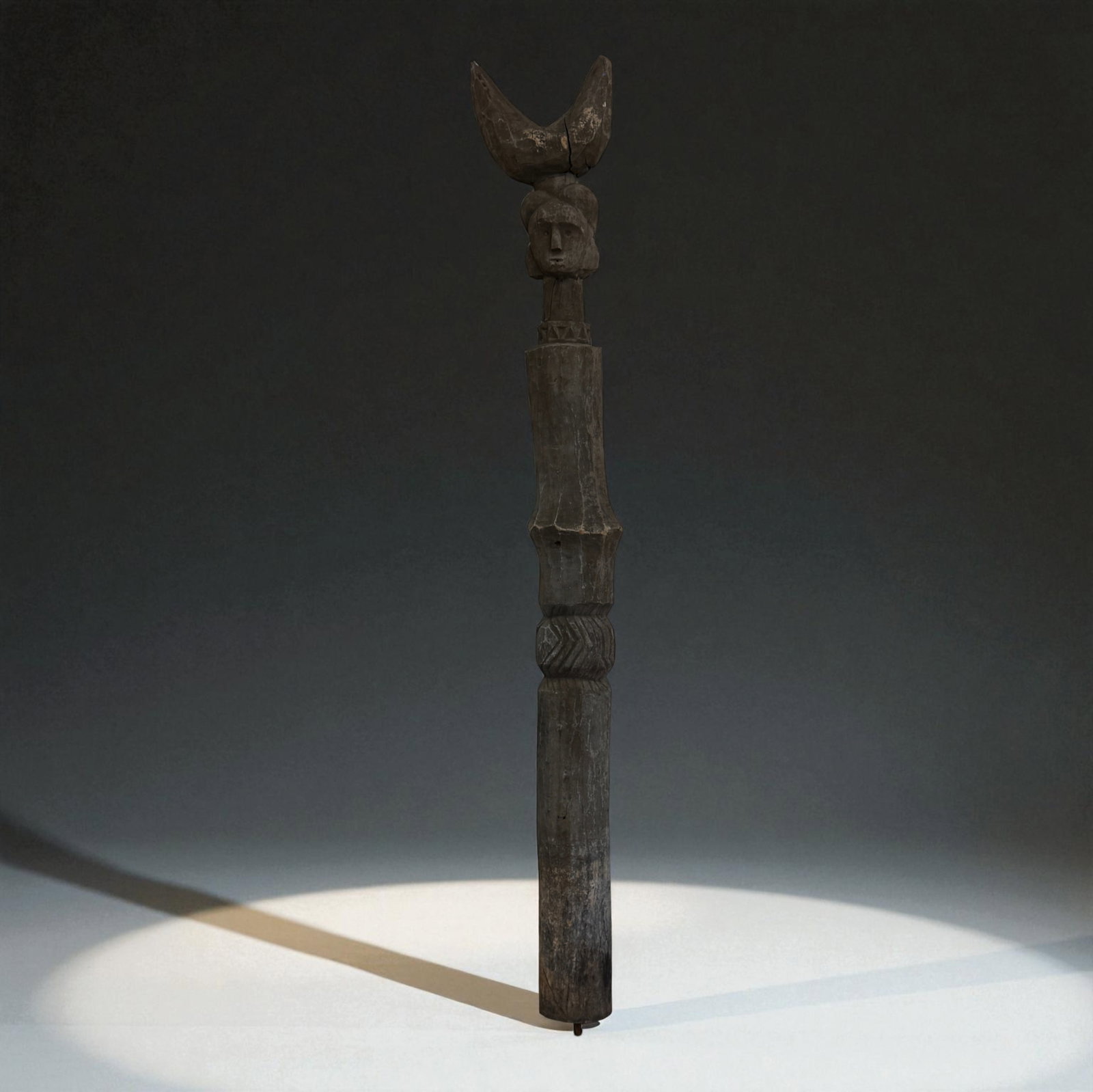 Large African Ritual Staff with Crescent Finial: Tall African carved wooden ritual staff topped by a crescent-shaped finial surmounting a human head, the shaft decorated with carved bands and varying profiles, retaining a rich, dark patina and signs