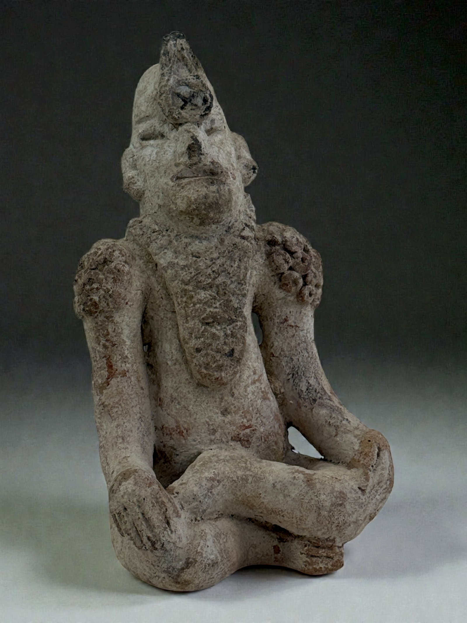 Seated Figure with Tall Headdress: Terracotta figure with tall headdress and folded legs, traces of red slip and burial patina; may represent a shaman or priestly figure. 6" tall.