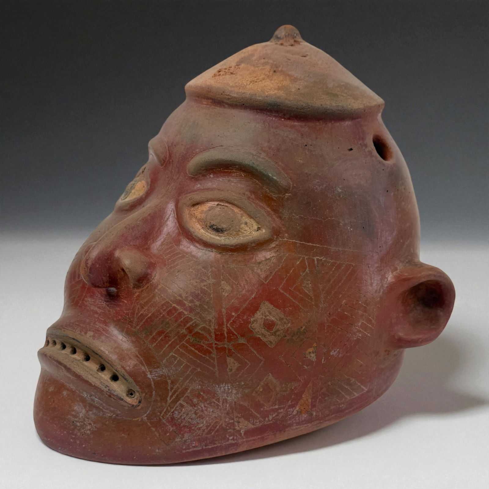 Pre-Columbian Large Red Head Effigy: Oversized terracotta head effigy with deeply carved features, incised facial motifs, and perforated teeth rows. Rich red slip surface with areas of earthen wear enhances its bold expression. Provenanc