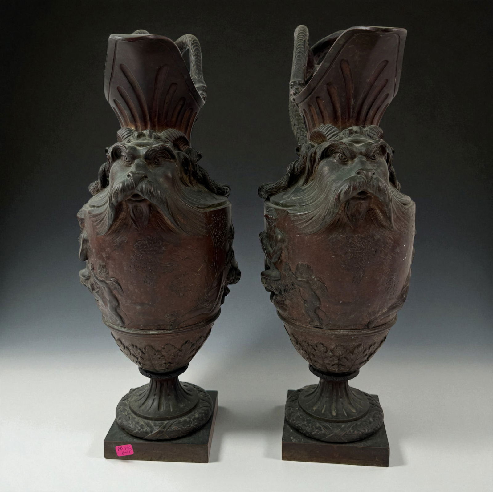 Antique Pair of Bronze Mythological Face Urns: Impressive pair of bronze urns featuring mythological bearded faces, foliate handles, and cherub relief scenes. Dark patina, 19th to early 20th century decorative bronzes. 19" inches