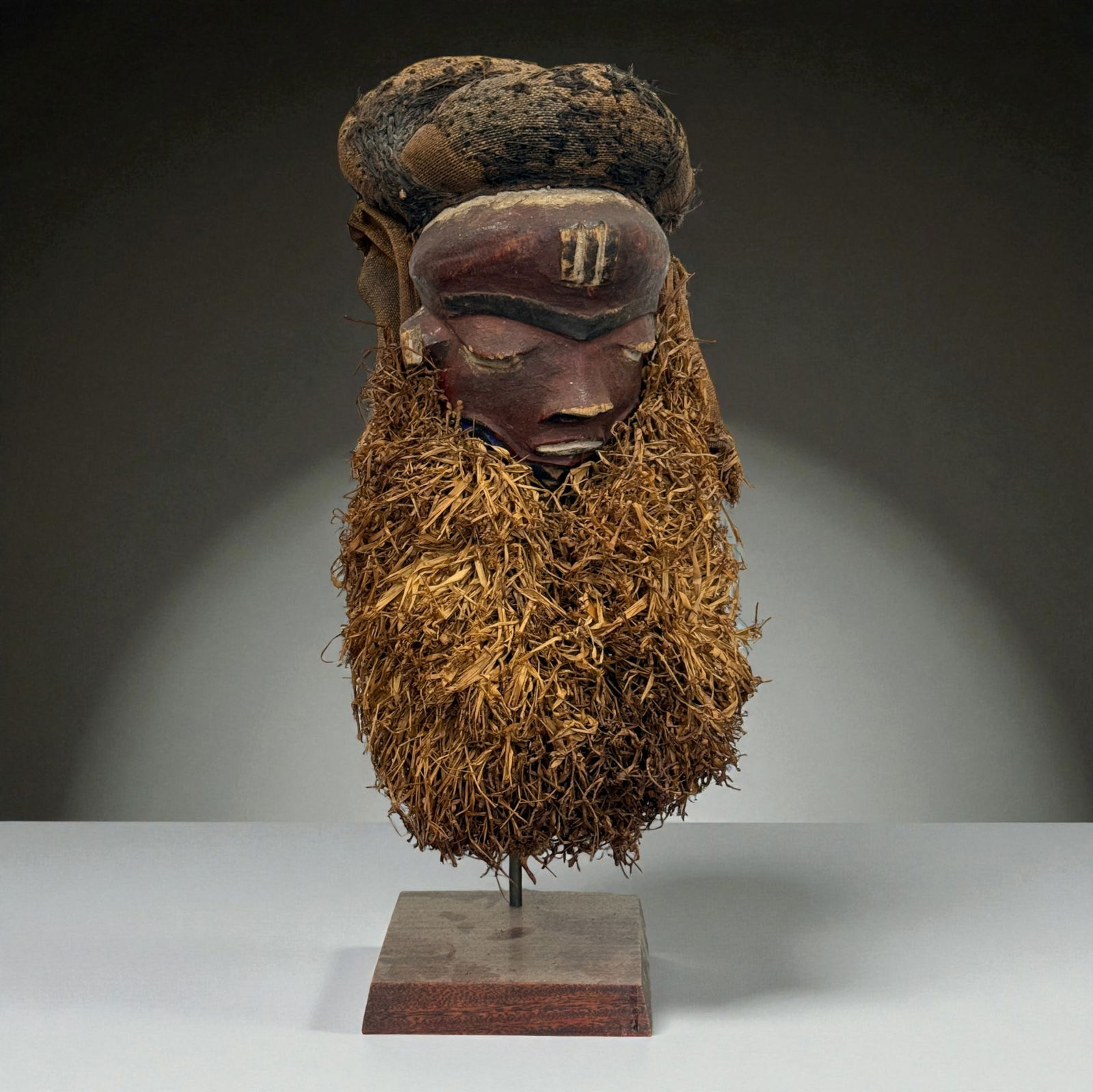 African Helmet Mask with Fiber Beard: Impressive carved wooden helmet mask with closed eyes, domed coiffure and dense fiber beard, mounted on a custom stand. Provenance: Beverly Hills Barakat Gallery Collection. 13"