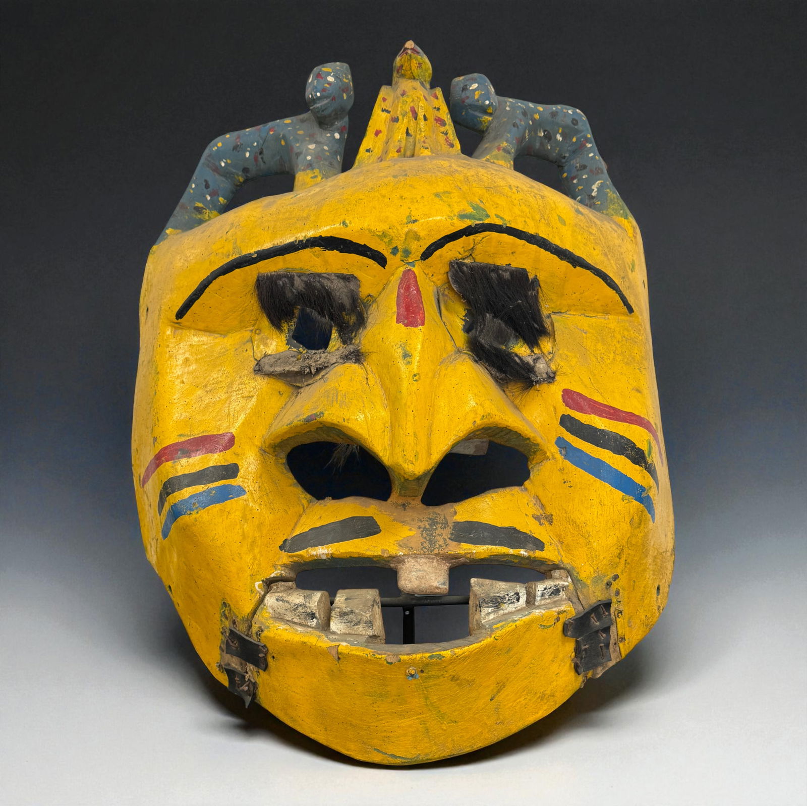 African Huge Polychrome Mask with Figures: Large polychrome African carved wooden mask with bold yellow ground, exaggerated nose and open mouth showing carved teeth, painted facial markings and applied hair brows, surmounted by two small seate