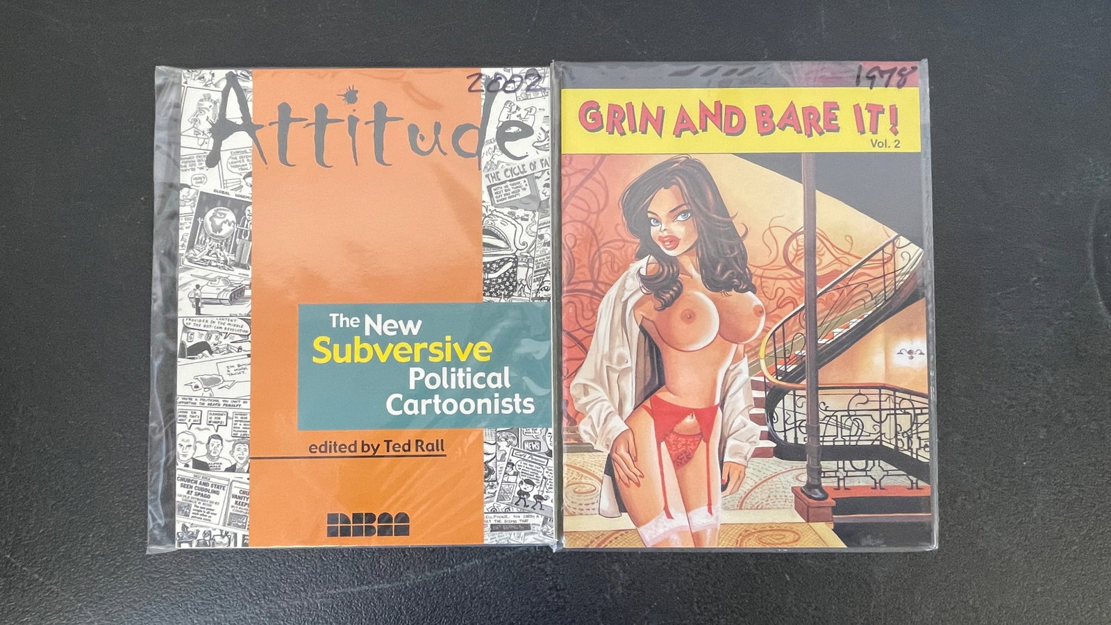Attitude: New Subversive Political Cartoonists (2002) & Grin and Bare It! Vol.2 (1978): A pairing of two adult-oriented cartoon compilation books, both sealed in protective plastic bags. The first is 'Attitude: The New Subversive Political Cartoonists' (2002), edited by Ted Rall and publ