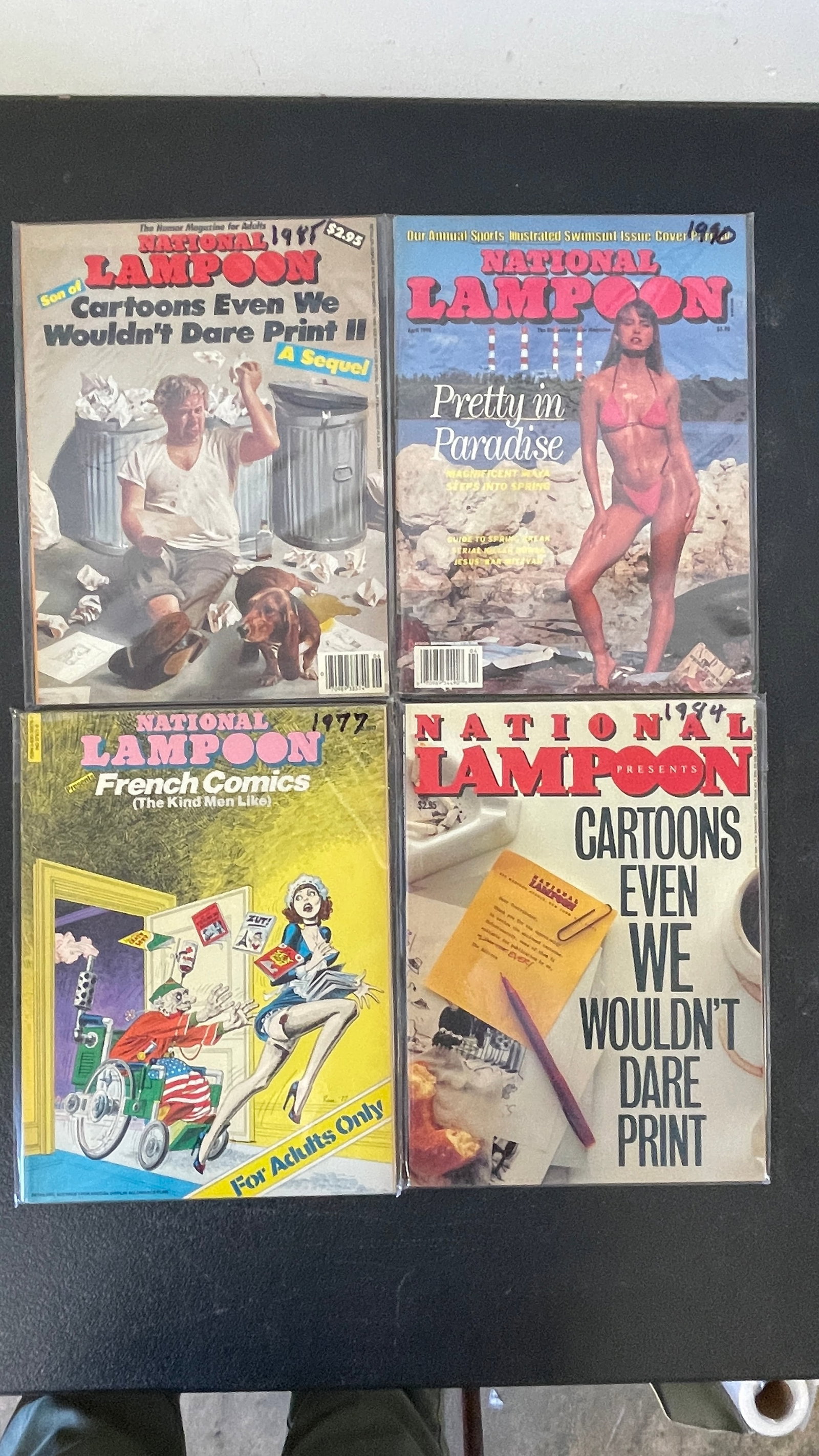 National Lampoon Magazine Lot x4 | 1977, 1984, 1985, 1990s Issues: A collection of four National Lampoon humor magazine issues spanning from 1977 to the mid-1990s, all stored in protective plastic sleeves. Titles include 'French Comics (The Kind Men Like)' (1977), 'C