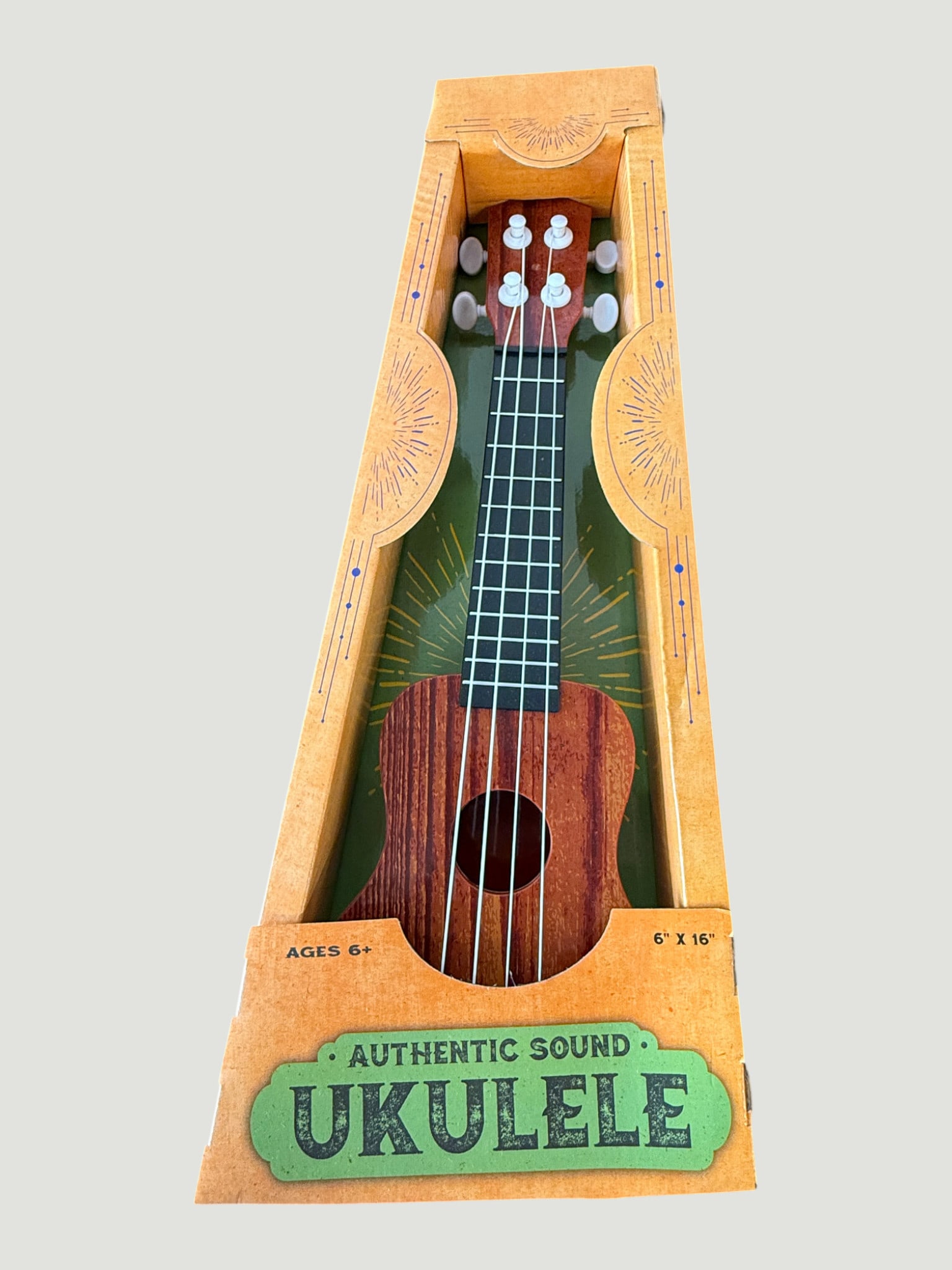 Authentic Sound Ukulele Toy, Ages 6+, 6"x16", Boxed: A soprano-style toy ukulele marketed as producing 'Authentic Sound,' presented in its original decorative cardboard display box with a green and gold sunburst graphic interior. The instrument