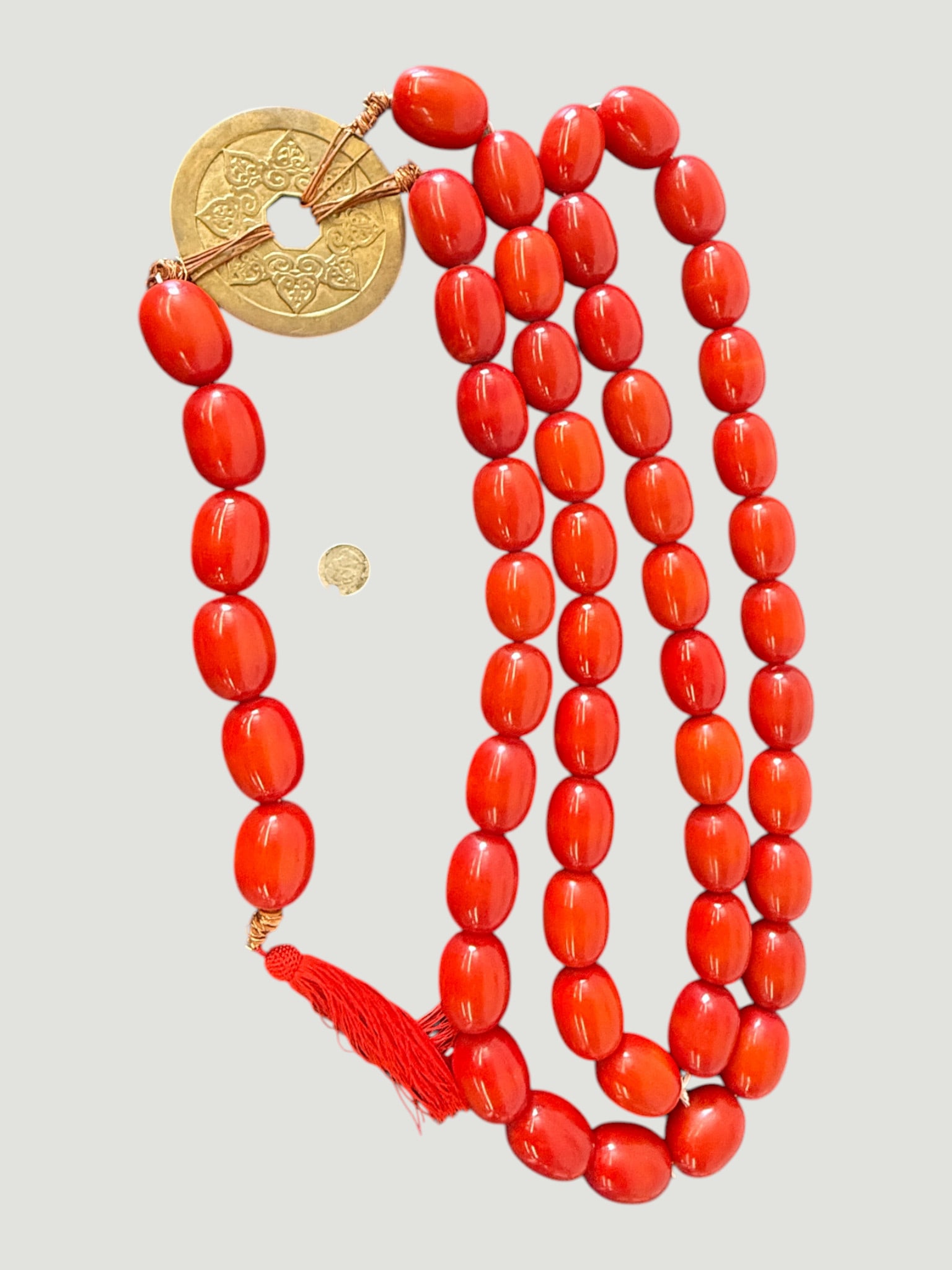 Red Coral-Style Oval Bead Mala Necklace with Gold-Tone Chinese Coin Pendant: A long mala-style prayer bead necklace featuring approximately 108 oval beads in a vivid red-orange coral color, likely composed of resin or dyed bone/synthetic material. The necklace is adorned with 
