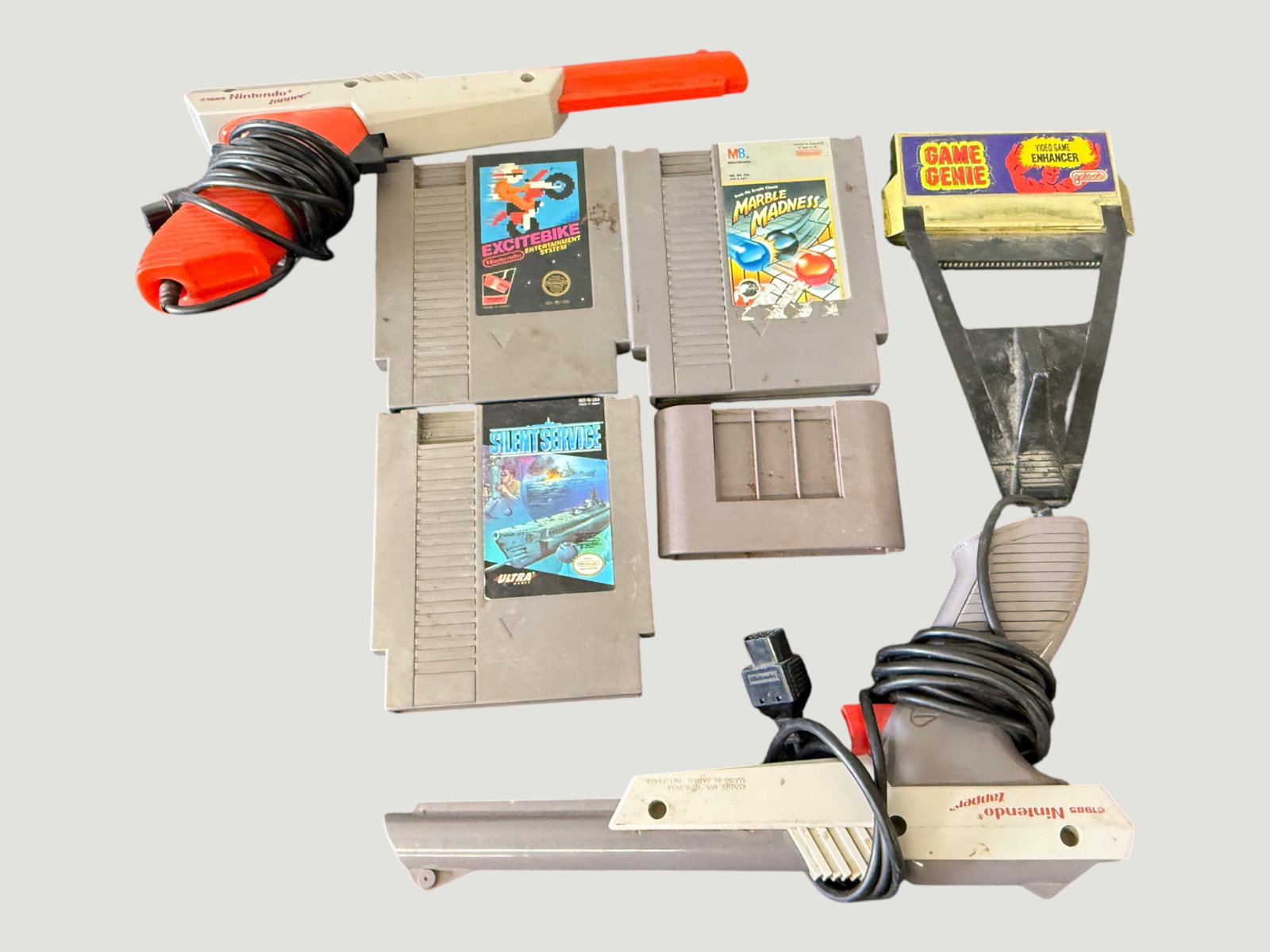 Nintendo NES Zapper Light Guns x2, Game Genie, & NES Game Cartridges Collection: A collection of Nintendo Entertainment System (NES) accessories and game cartridges from the mid-to-late 1980s. Includes two NES Zapper light guns (one orange/red model circa 1985, one grey model circ
